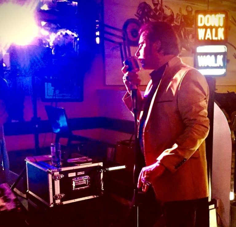 Alleinunterhalter Elvis Ein Alleinunterhalter im Elvis Outfit singt in ein Mikrofon vor bunten Lichtern und einem „Don't Walk“-Schild.