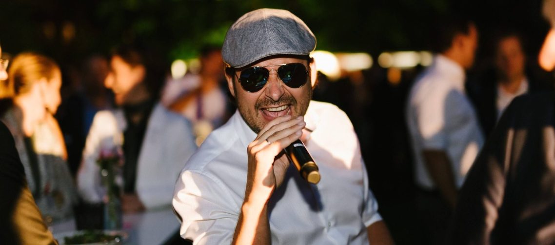 Adriano Celentano Cover Sänger Mann mit Sonnenbrille und Kappe singt als Adriano Celentano Imitator in Mikrofon, umgeben von feiernden Gästen.
