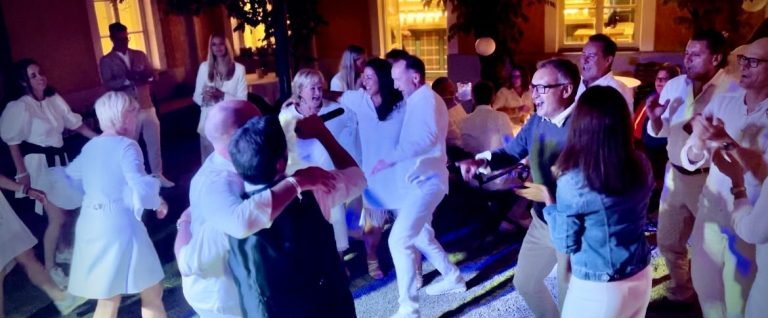 Menschen in weißen Outfits tanzen gemeinsam mit Roland Kaiser Double Sänger bei einer Feier, mit bunten Lichtern im Hintergrund.