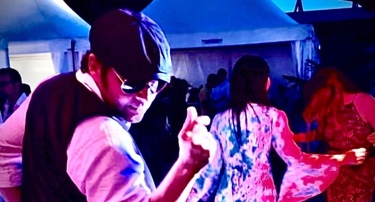 Italienische Showeinlage Mann in Sonnenbrille und Italo Outfit tanzt, umgeben von festlich gekleideten Personen.