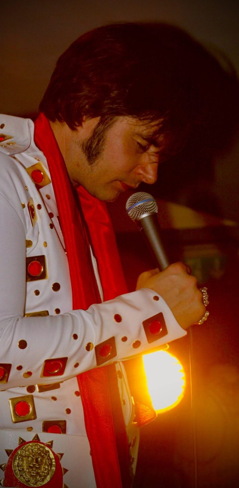 Elvis Imitator Österreich Mann als Elvis Double mit Mikrofon, in einem Elvis-inspirierten Outfit und rotem Schal.