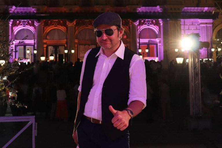 Italo Sänger buchen Mann in Sonnenbrille und Weste steht als Italo Sänger vor bunt angestrahltem Gründerzeitgebäude bei Nacht.