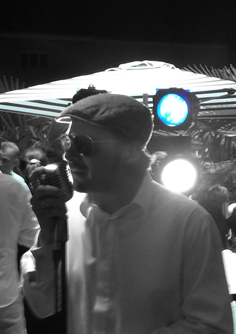 Italienischer Sänger Österreich Mann mit Hut und Sonnenbrille singt ins Mikrofon, umgeben von Bühnenlichtern.