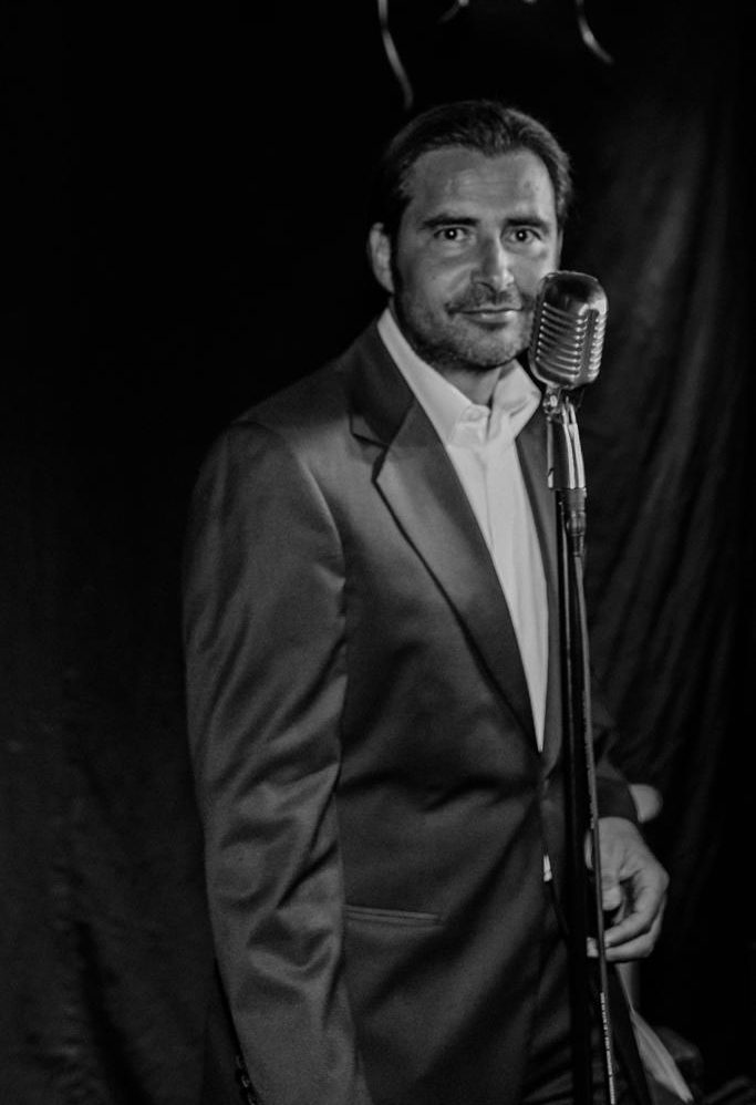 Mann im Smokingsakko steht an einem Mikrofon mit schwarzem Hintergrund.