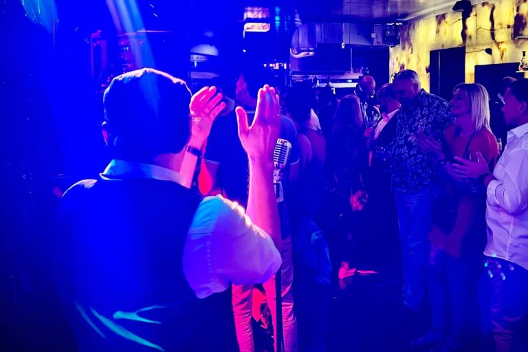 Italo One Man Show Sänger steht vor Menschen die tanzen am Mikrofon in einer lebhaften Bar mit bunten Lichtern und Musik.