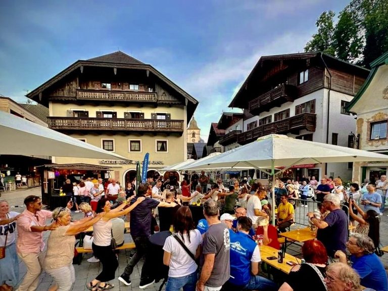 Menschen feiern bei einem Austropop Abend und tanzen auf einer Straße mit Zelten, Bänken und traditionellen Gebäuden im Hintergrund.