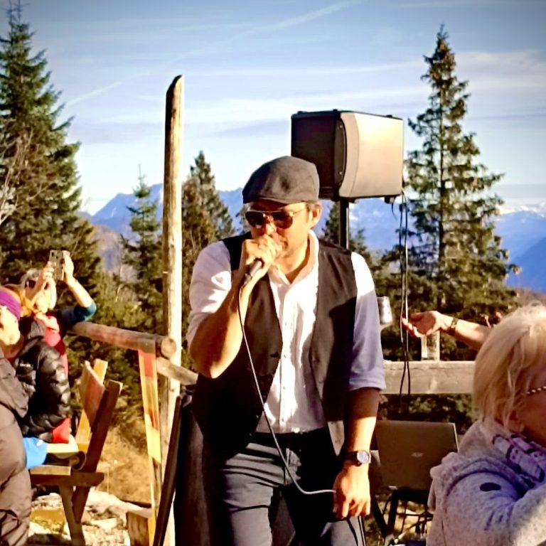 Mann singt als Austropop Sänger mit Mikrofon und Lautsprecher vor einer alpinen Kulisse aus Bäumen und Bergen.