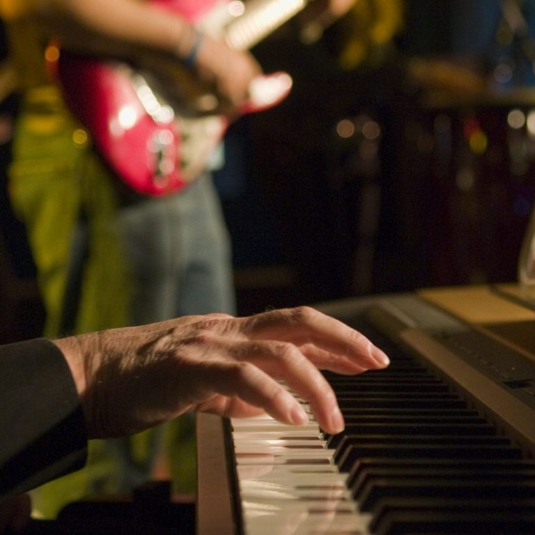 Hand spielt Klavier, im Hintergrund ein Musiker mit roter Gitarre.