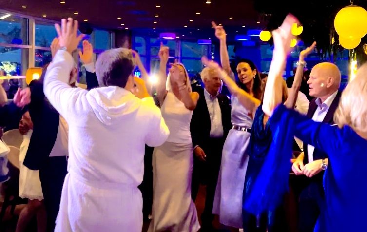 Menschen tanzen voller Begeisterung gemeinsam mit Udo Jürgens Imitator im Bademantel auf einer Feier, einige in formeller Kleidung.