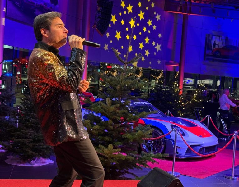 Schlagersänger buchen Mann singt auf einer Bühne mit Weihnachtsbaum und Sportwagen im Hintergrund.